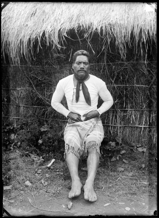 Unidentified Maori man beside a thatched whare, Te Kauri, Otorohanga District