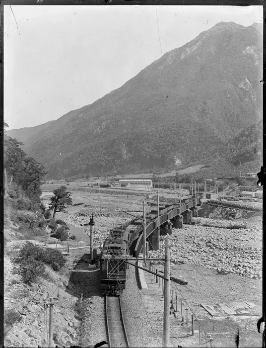 Locomotive towing wagons over a bridge and river foreground, rail yards, station and buildings below bush covered mountain beyond, possibly Otira, Westland