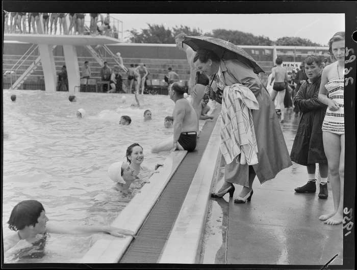 An unidentified woman, sheltering under an umbrella and talking to swimmers, Naenae Olympic Swimming Pool, Lower Hutt