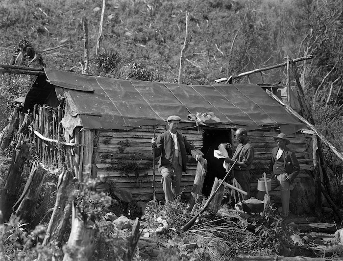 A scrubland scene showing a hut with three men standing outside
