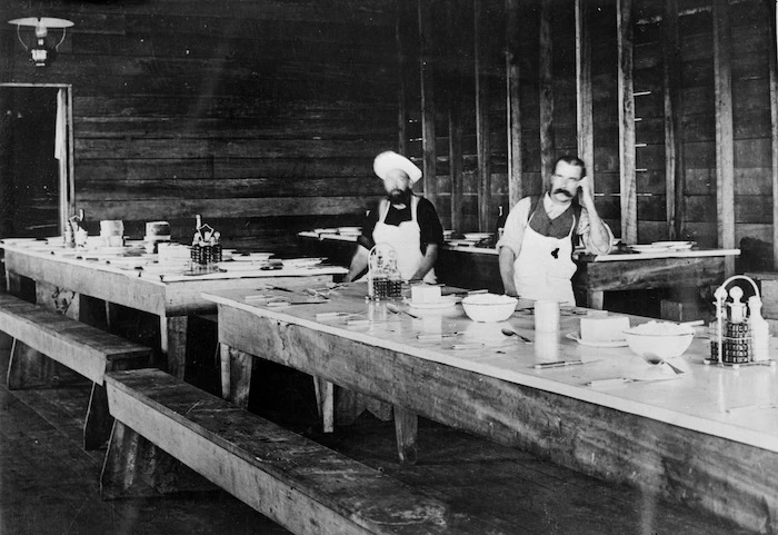 Two cooks posing by tables set for a meal, in a timber camp dining room, Mokai