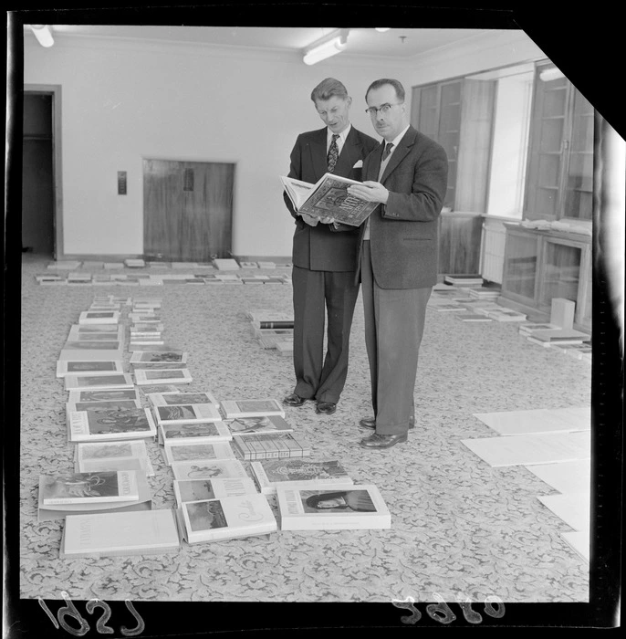 Tony Murray-Oliver with an unidentified man looking at a book with other books for the Alexander Turnbull Library laid out on the floor, Wellington